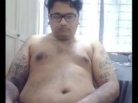 Vaibhav, un indio fijiano peludo, se sienta desnudo en una silla de oficina negra en su dormitorio, con su barriga y tatuajes visibles. Se masturba, sus manos regordetas trabajando sobre su miembro flácido, apuntando a un clímax explosivo.
